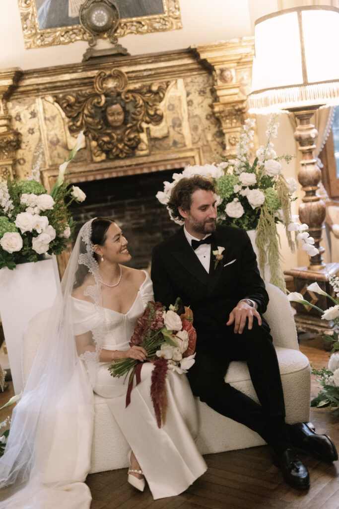 Novios sentados junto a la chimenea barroca de La Baronía rodeados de decoración floral en blanco: calas, rosas y hortensias diseñadas por 18 Pétalos