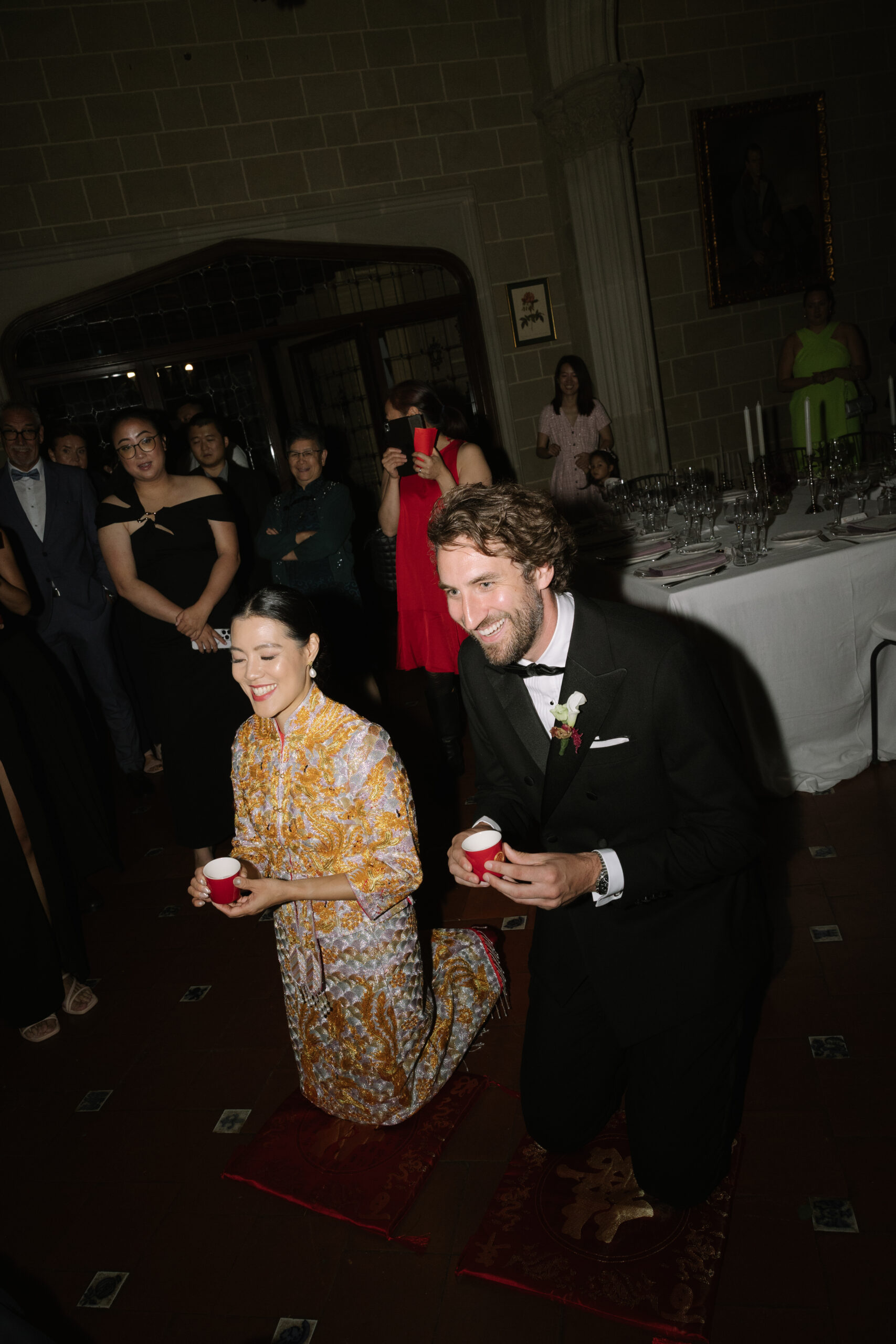 Pareja realizando la ceremonia del té china en La Baronía: novia con qipao dorado arrodillada junto al novio con esmoquin, sosteniendo tazas rojas de la tradición nupcial china